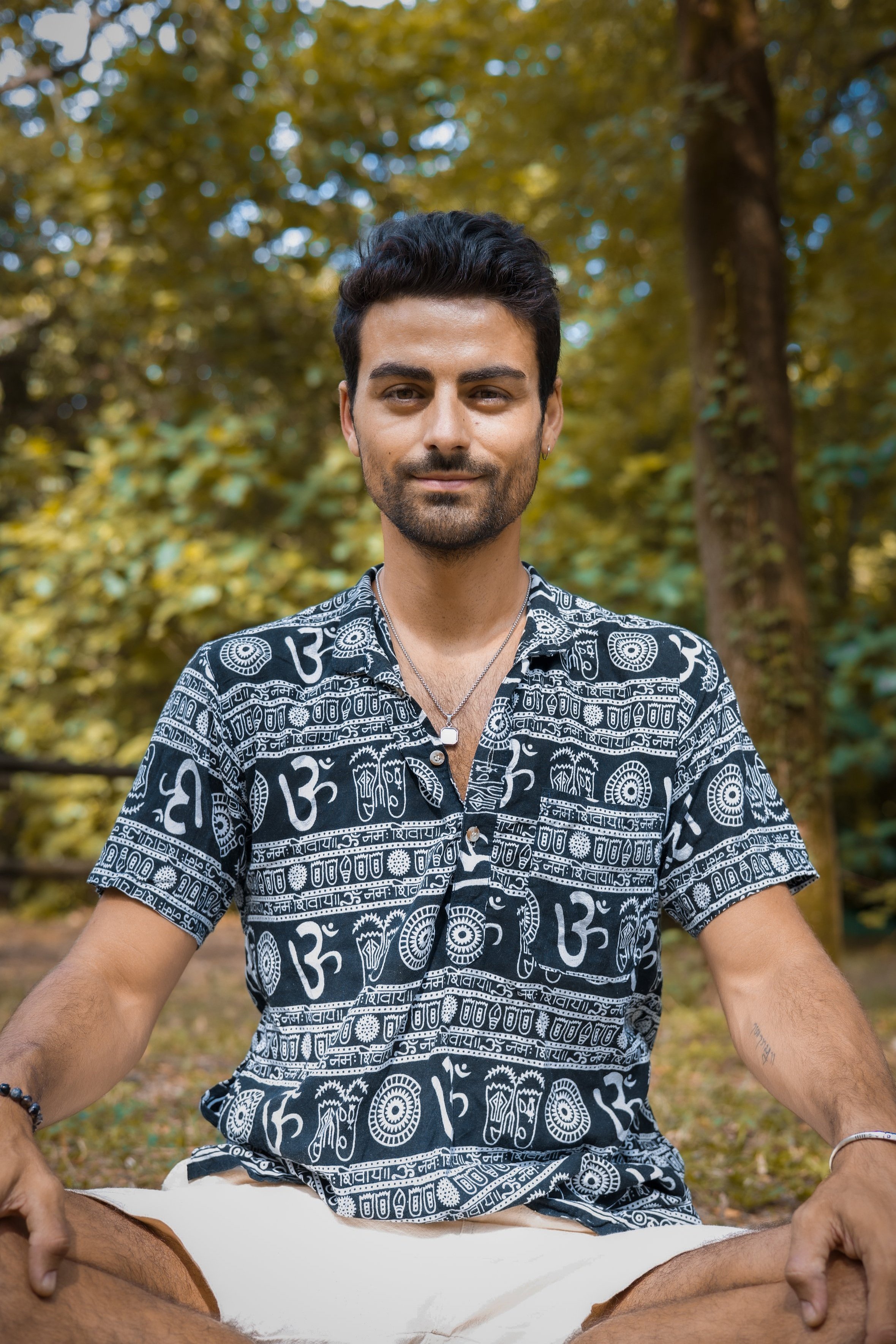 Un homme assis en position de méditation dans un parc avec des arbres verts en arrière-plan, portant une chemise à motifs et des bijoux.