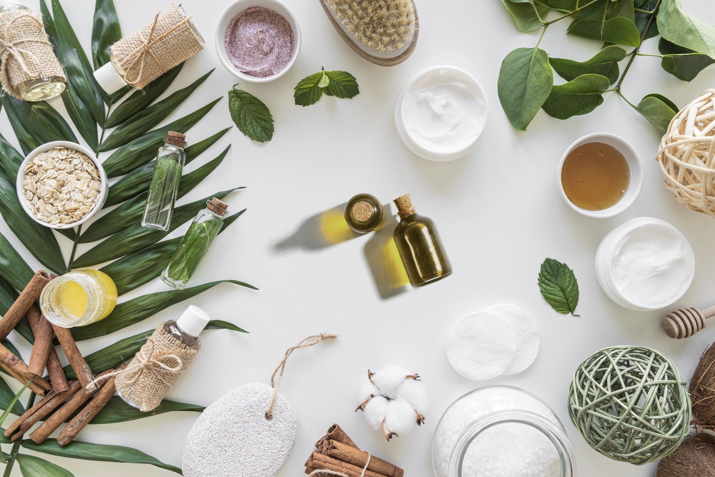 Produits de soins naturels sur une surface blanche, incluant des huiles essentielles, des crèmes, des feuilles vertes, des bâtons de cannelle, des boules de coton, des écorces, et des objets en matériaux naturels.